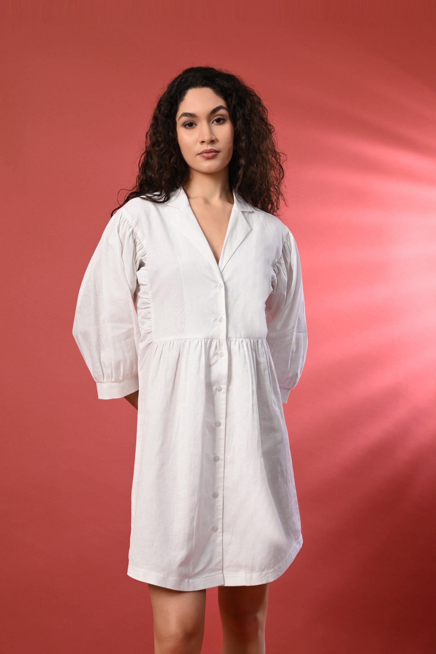 A white V-neck linen shirt dress with a relaxed fit, standing in a bright, summery setting. The lightweight fabric 