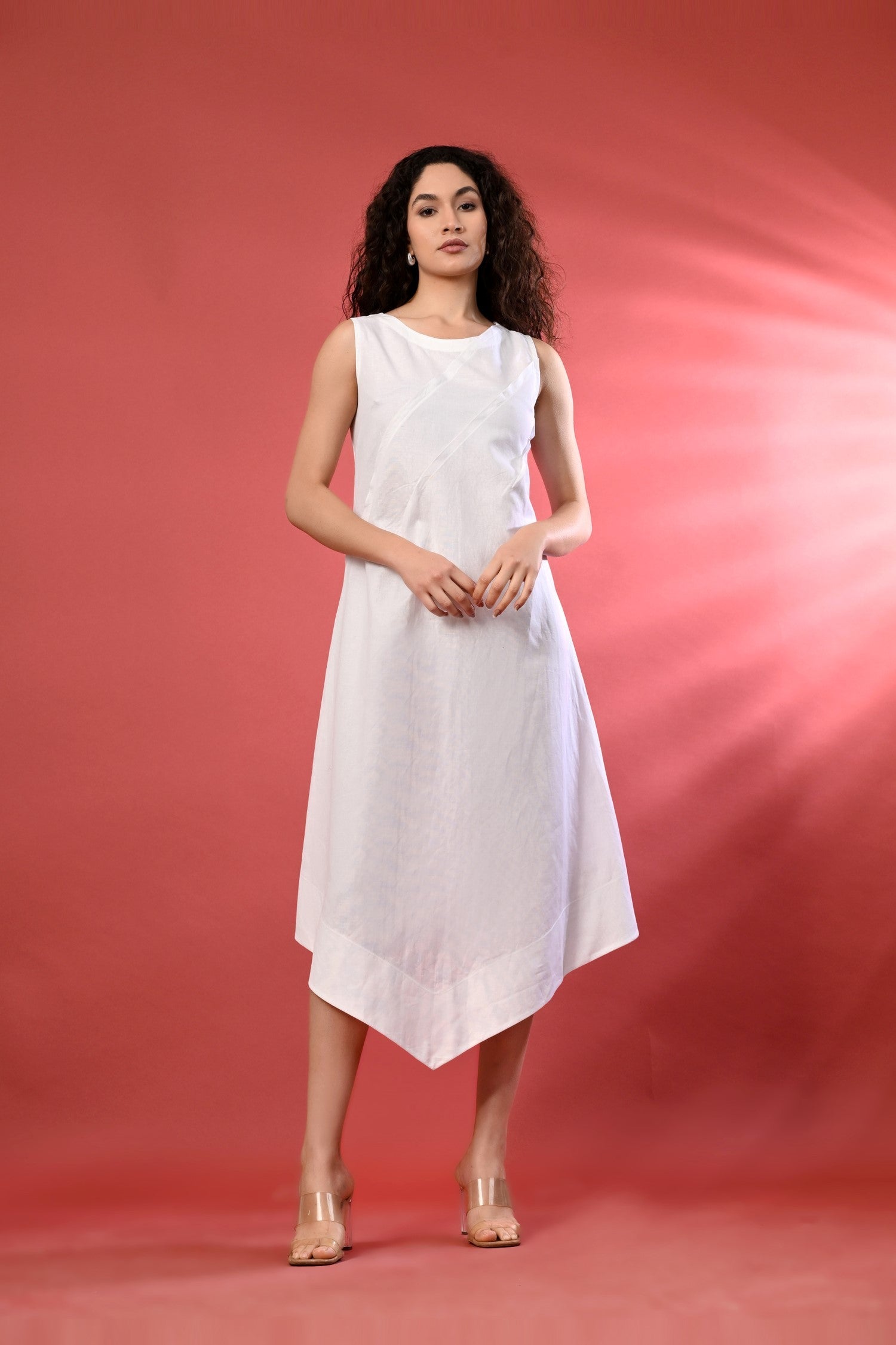 White sleeveless linen dress with a fit-and-flare silhouette and high-low hem, standing in a sunlit outdoor setting. 